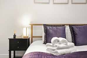 A neatly made bed with a purple blanket and pillows, a bedside table with a lamp and a book, and a small vase with reeds. - 23A Bevenden Street (Greater London)