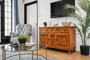 A living room with a tufted armchair, a wooden cabinet, a glass coffee table, and a potted plant.