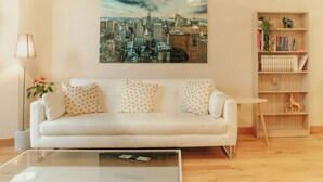 Un salon moderne avec un canapé blanc, une table basse en verre, une bibliothèque et un tableau représentant un paysage urbain accroché au mur.