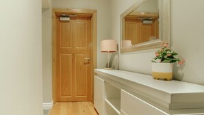 Un couloir avec une porte en bois, une console blanche surmontée d’un miroir, une lampe et une plante en pot.