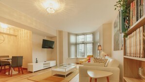 Un salon moderne avec un canapé blanc, une table basse et un téléviseur fixé au mur. Une bibliothèque remplie de livres et une plante se trouvent sur le côté droit.