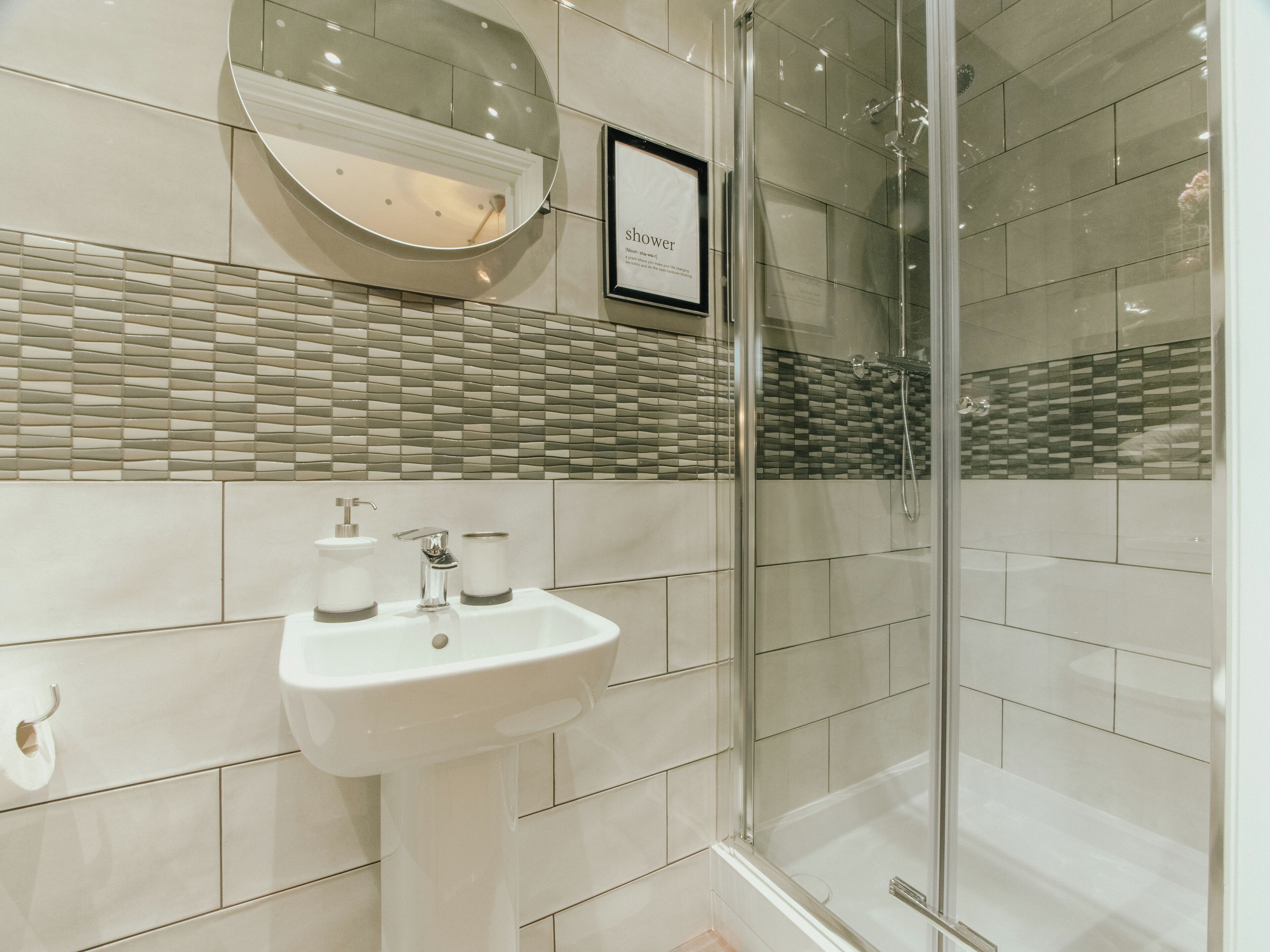 Une salle de bain avec une douche à parois de verre, un lavabo blanc doté de deux distributeurs de savon et un panneau encadré placé au-dessus du miroir.