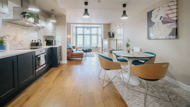 A modern kitchen with dark cabinets, a marble countertop, and a built-in oven. There is a dining area with a table and chairs, and a large window providing natural light. - Stunning Quiet Apartment in Historic Building (Sheffield)
