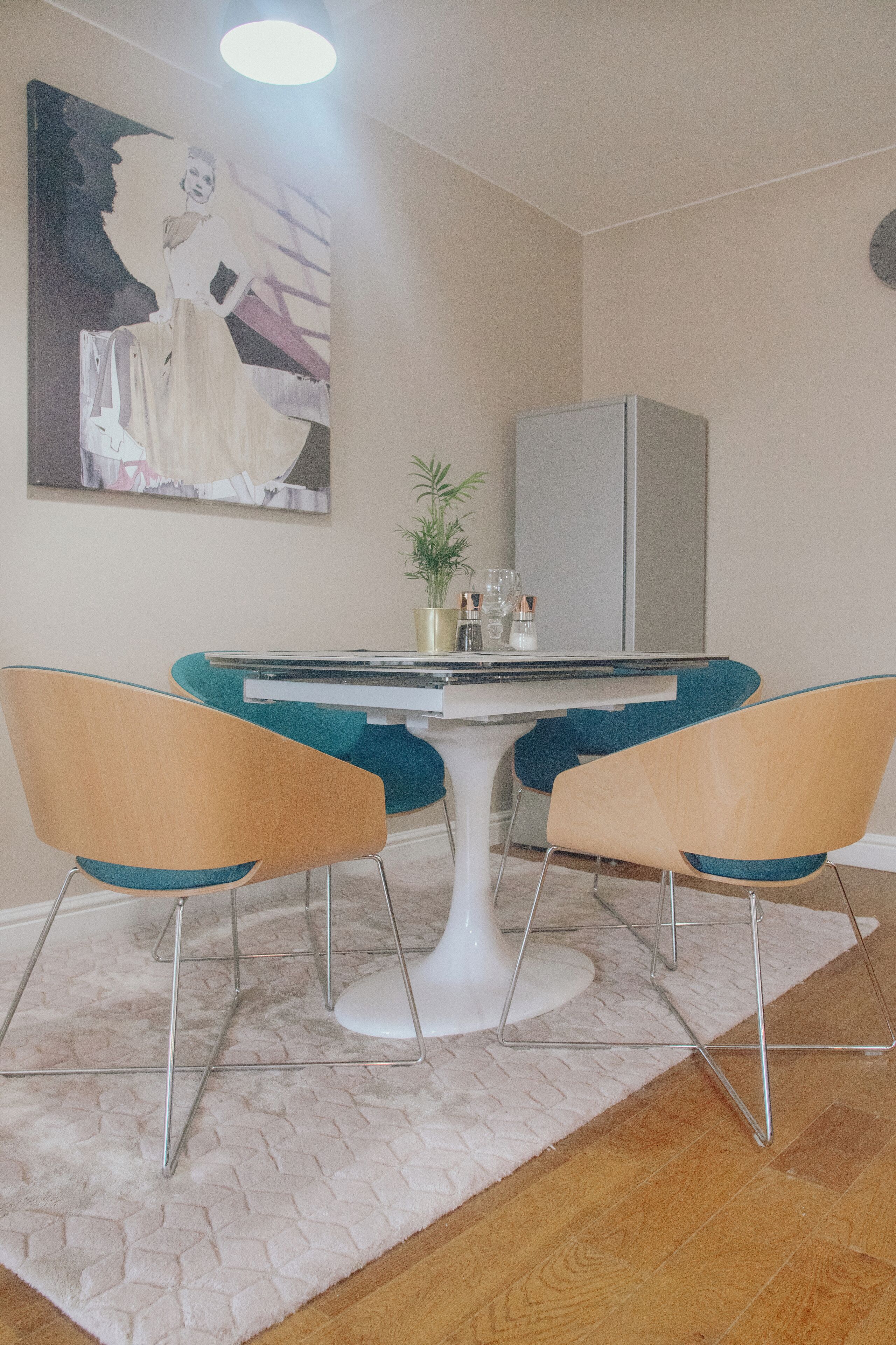 Un comedor con una mesa blanca, sillas color turquesa, un florero blanco con una planta y un refrigerador blanco.