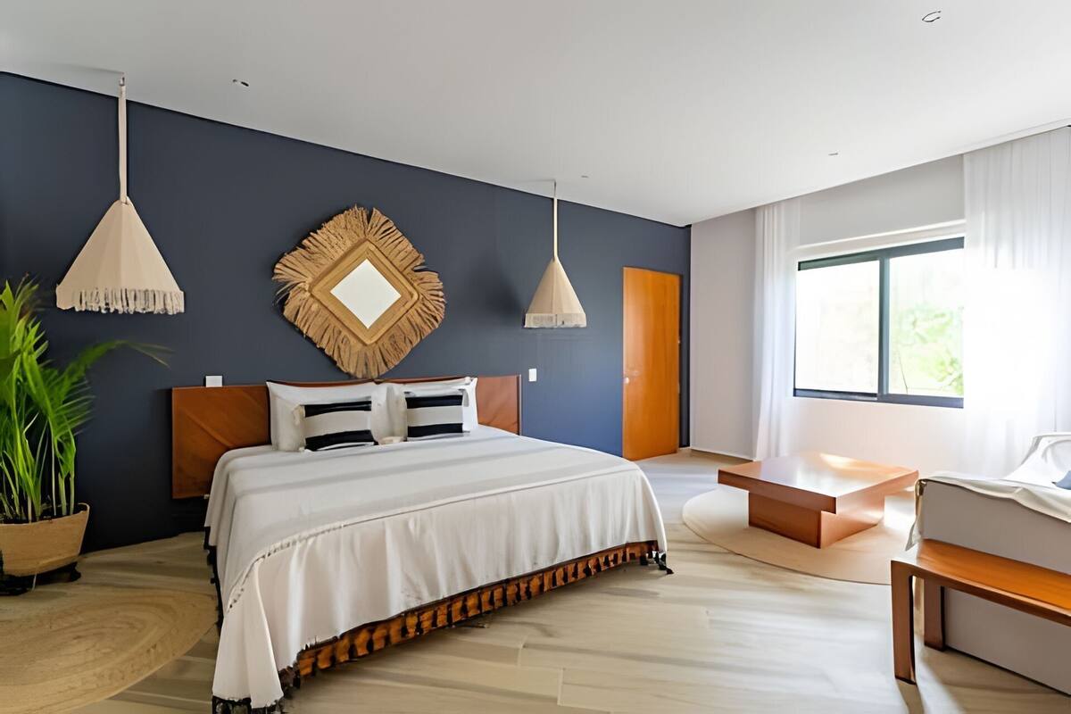 A modern bedroom with a large bed, two pendant lights, a wooden headboard, a large mirror, a wooden bench, and a large window with curtains.