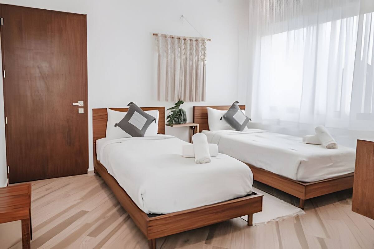 A hotel room with two wooden beds, white bedding, a wooden nightstand, a potted plant, and a large window with sheer curtains.