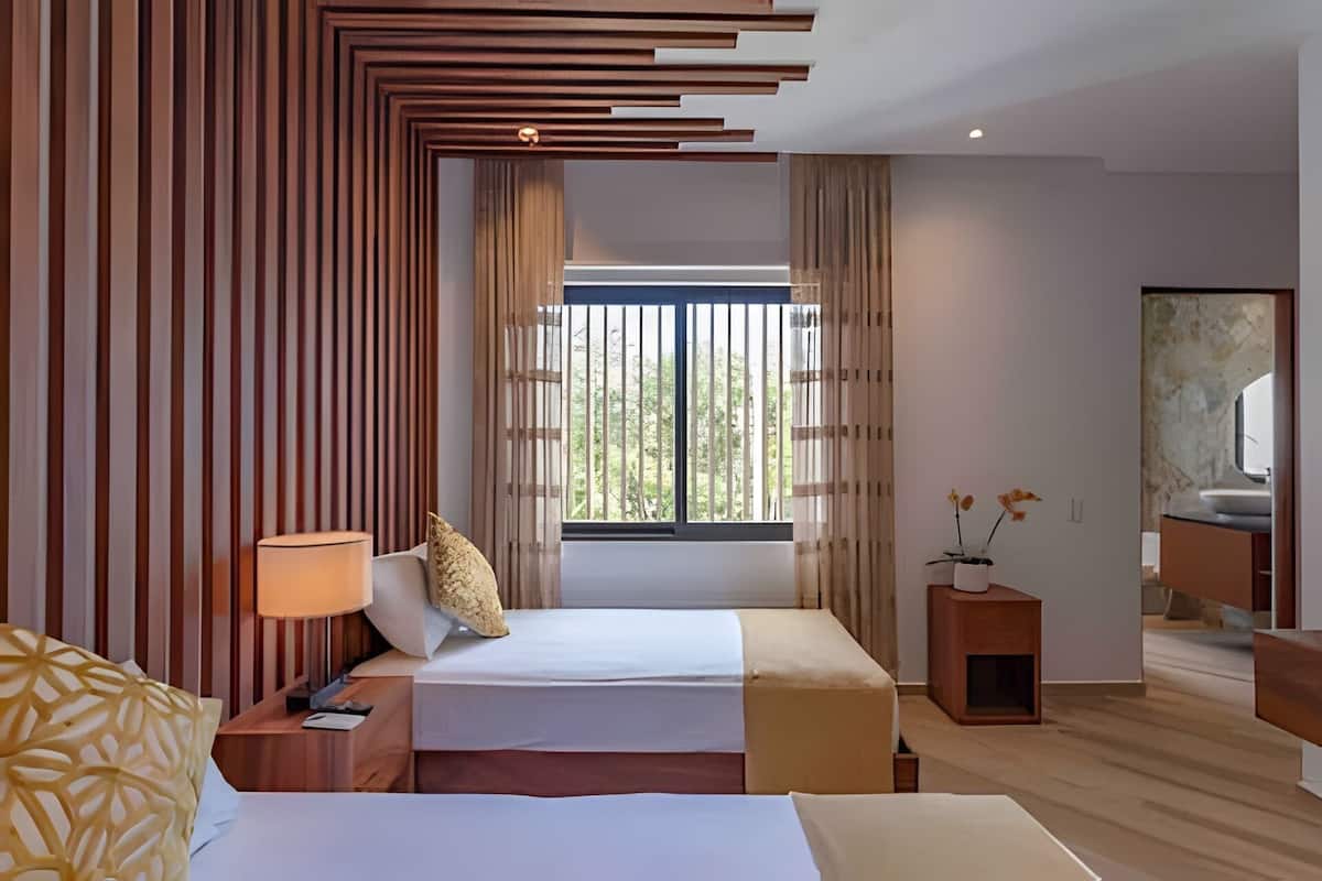 A hotel room with two beds, a bedside table with a lamp, a small wooden side table, a window with curtains, and a view of greenery outside.