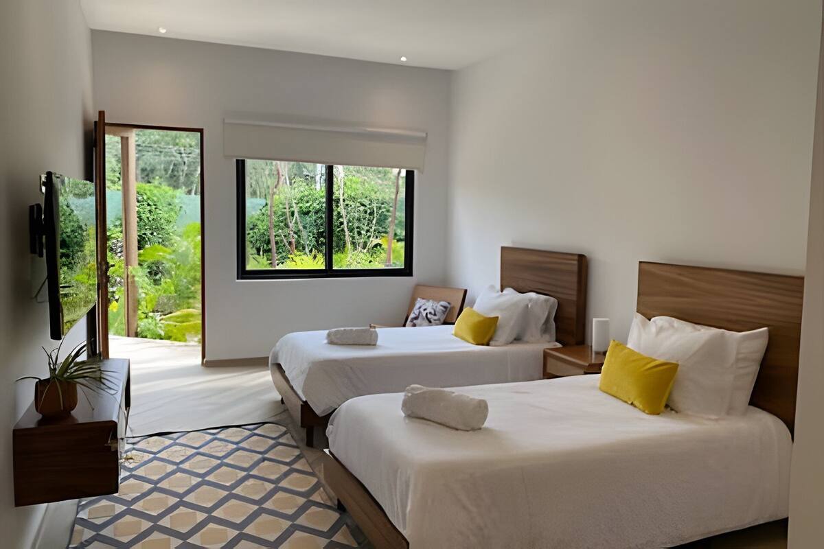 A hotel room with two beds, a large window with a view of greenery, a TV, and a small plant.