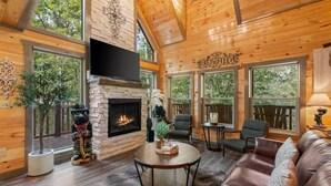 A cozy cabin interior with a stone fireplace, leather furniture, and large windows offering a view of the outdoors. - INDOOR HEATED POOL CABIN SLEEPS 10 & GAME LOFT “BEAR SPLASH” NEAR PARKWAY! (Sevierville)
