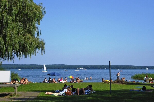Une scène au bord d’un lac, avec des personnes se relaxant sur l’herbe, des bateaux sur l’eau et un voilier à l’horizon.