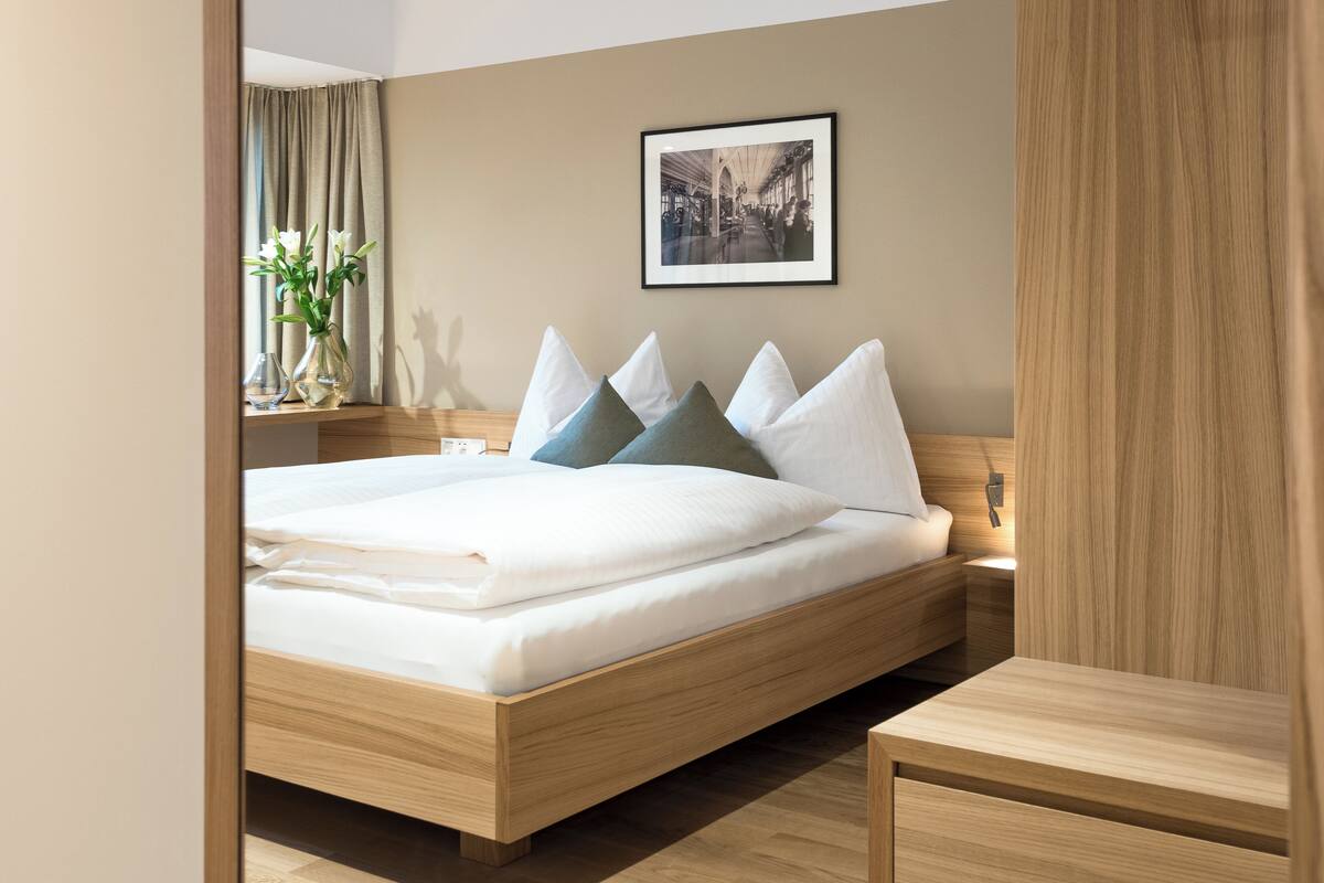 A modern hotel room with a wooden bed, a nightstand, a bedside table, a framed picture on the wall, and a window with curtains.