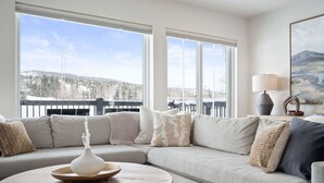 A modern living room with a large sectional sofa, a round wooden coffee table, and a view of a snowy landscape through large windows. - Modern Mountain Retreat (Silverthorne)