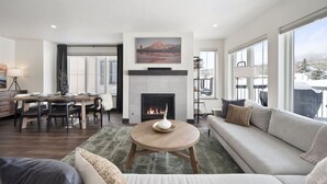 A modern living room with a fireplace, a dining area, and a large window offering a view of the outdoors. - Modern Mountain Retreat (Silverthorne)