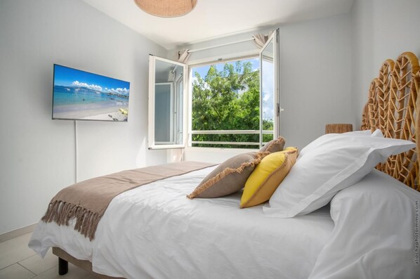 A bedroom with a bed, a TV, a window with an open view, and a wicker headboard. - Horizon Bleu "feet in the water (Les Trois Ilets)