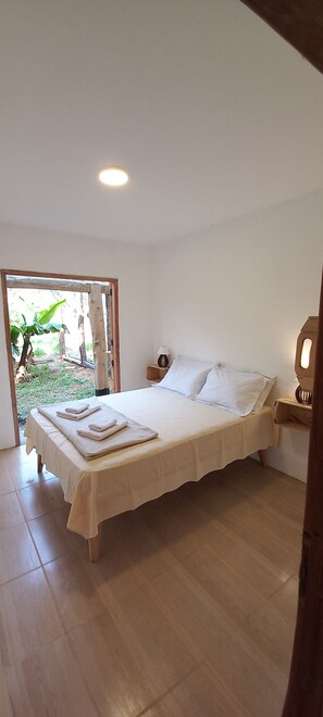 Un dormitorio con una cama grande, una mesita de noche y vistas a un jardín a través de una puerta abierta.