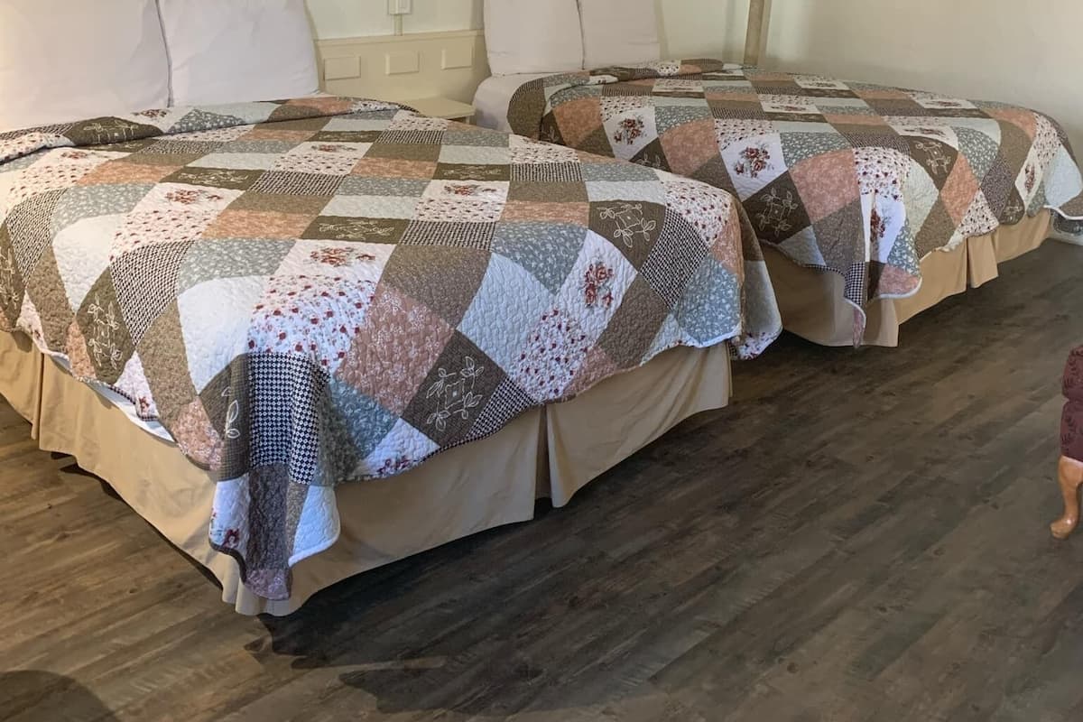 A hotel room with two beds, each covered with a patchwork quilt. There is a framed picture on the wall above the beds, a bedside lamp, and a small table between the beds.