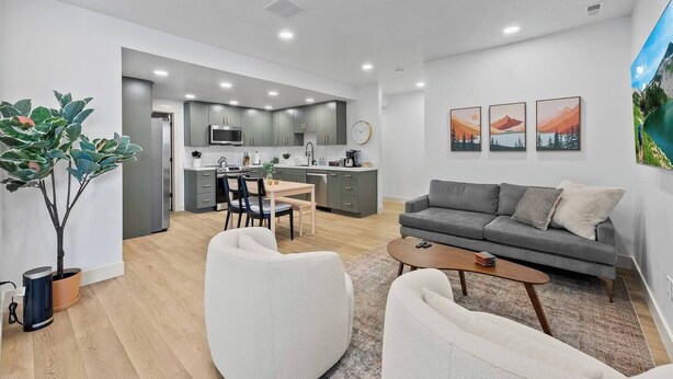 Ein modernes Wohnzimmer mit einem grauen Sofa, weißen Sessel und einem Couchtisch. Es gibt einen Essbereich mit Tisch und Stühlen sowie eine Küche im Hintergrund.