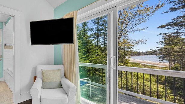 A room with a white armchair, a flat-screen TV, and a large sliding glass door leading to a balcony with a view of trees and a beach. - Secluded Cottage with Beach Access! [Cove Cottage] (Tremont)
