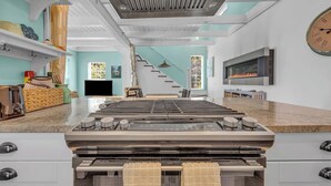 A modern kitchen with a large stove, a dining area with a high table, and a staircase leading to an upper level. - Secluded Cottage with Beach Access! [Cove Cottage] (Tremont)