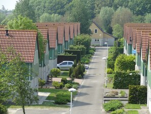 Egy lakóövezet, ahol terrakotta színű cserépfedeles házak, zöld zsalugáterek és egy központi út található.