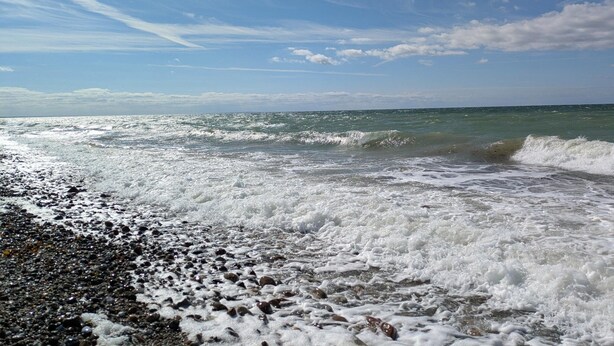 A beach with waves crashing onto a pebbly shore under a clear sky. - Ferienhaus in Bojendorf mit Terrasse (Fehmarn)