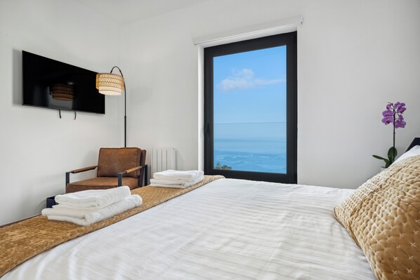 A bedroom with a large bed, a chair, a TV mounted on the wall, and a view of the ocean through a large window. - Casa Afonso (Funchal)