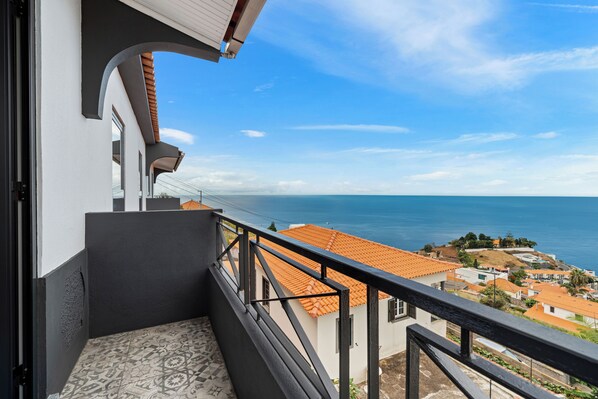 Un balcón con suelo de baldosas, barandilla y vistas al mar y edificios.