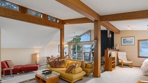 Un salon spacieux avec un plafond haut et des poutres en bois, un canapé jaune, un fauteuil rouge et une table basse en verre.