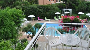 Un espace aménagé au bord de la piscine, avec des chaises et des parasols en plastique blancs, entouré d’une végétation luxuriante et avec un bâtiment visible au loin.
