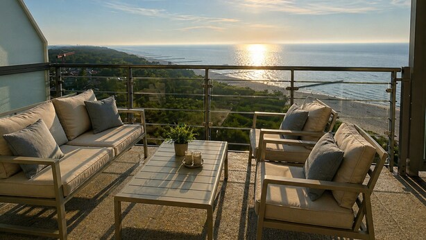 A balcony with a sofa, chairs, and a table, overlooking a coastal view with the sun setting over the ocean. - Apartment in Dziwnowek With Sea View (Dziwnów)