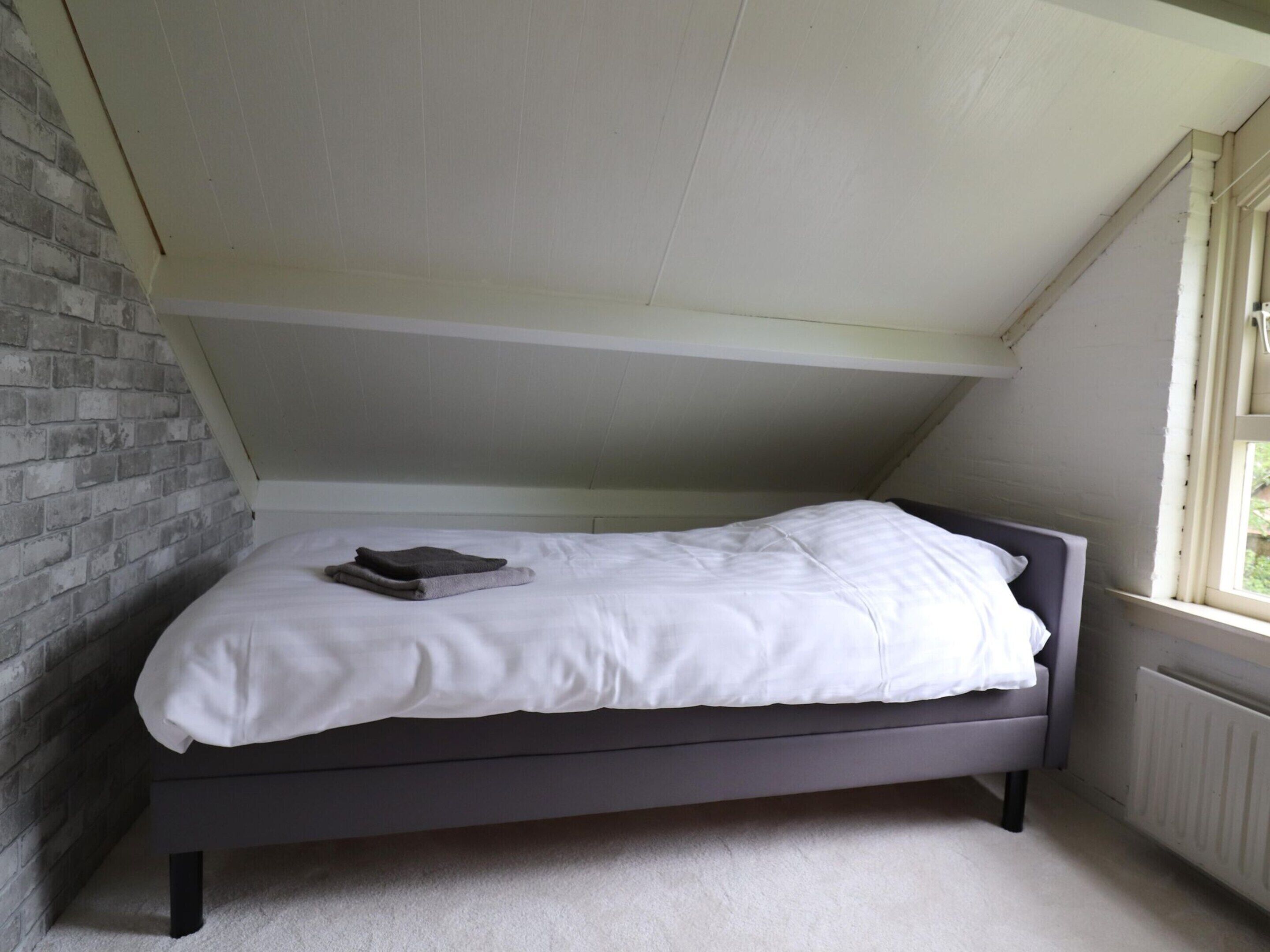 A single bed in a room with sloped ceilings, a brick wall on one side, and a window on the other.