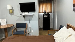 A hotel room with a bed, a desk with a lamp, a television, a kitchenette, and a chair.