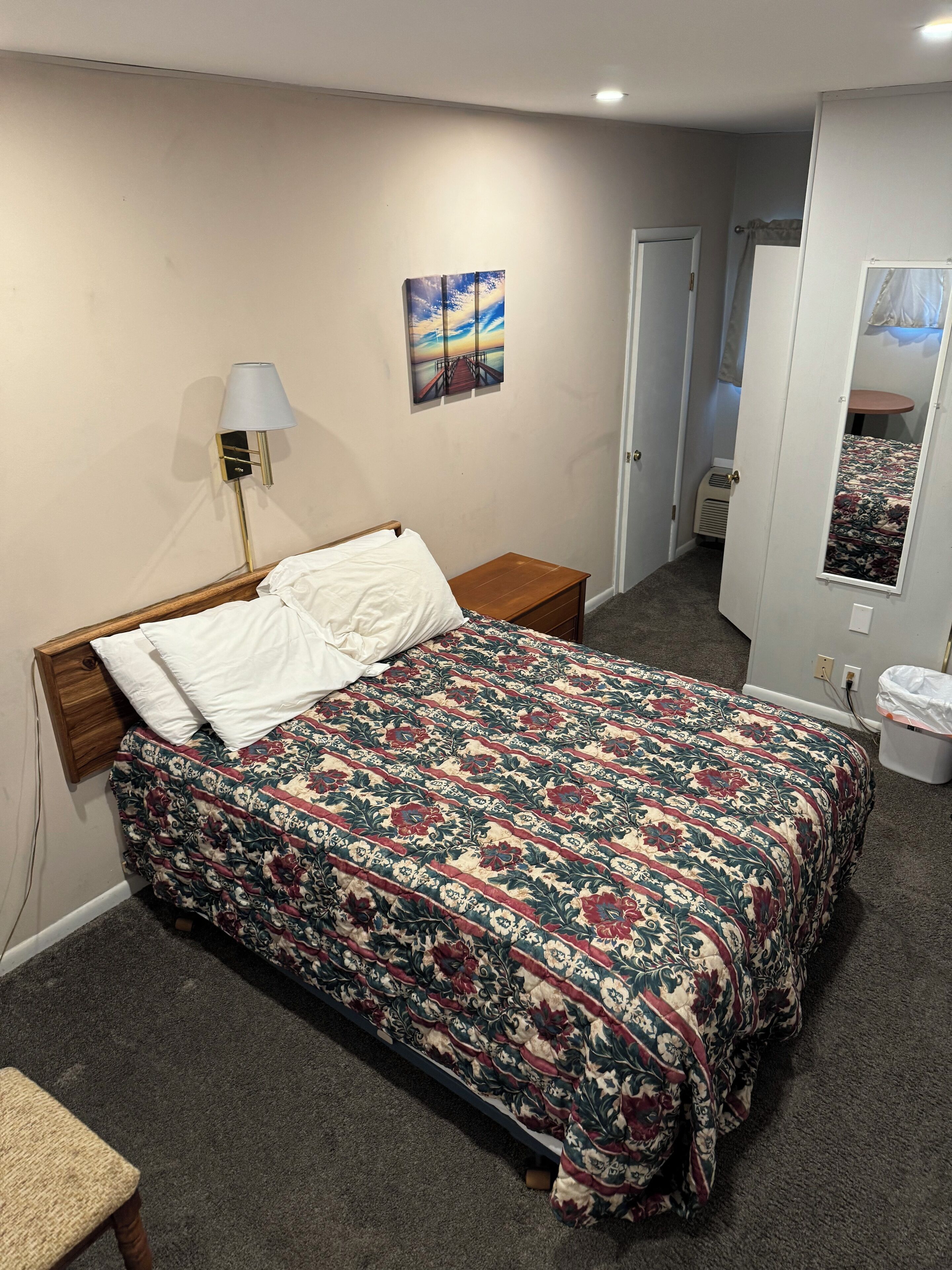 Een slaapkamer met een bed, een nachtkastje, een lamp, een deur, een spiegel en een tapijtvloer.
