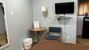 A hotel room with a large bed, a desk with a lamp, a chair, a TV mounted on the wall, and a small kitchenette area.