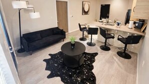 A modern living room with a black sofa, a round coffee table, and a black leather rug. There is a dining area with a bar and two bar stools. - The Luxe Black Haven (340) (Des Moines)