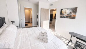 A bedroom with a large bed, a desk, a painting, and a laundry area. - The Luxe Black Haven (340) (Des Moines)