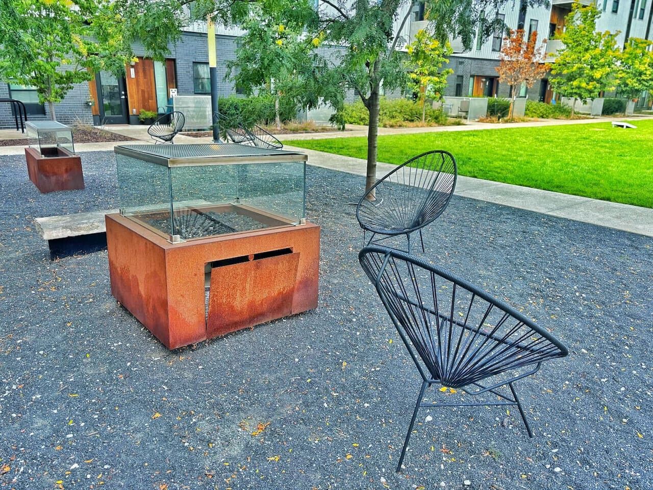 An outdoor seating area with a glass-top table and metal chairs, surrounded by a gravel surface and greenery.