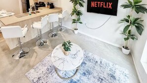 Una sala de estar moderna con una cocina al fondo, un sofá blanco, una mesa de centro y un televisor montado en la pared mostrando 'NETFLIX'.