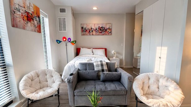 A modern bedroom with a grey sofa, two fluffy chairs, a glass coffee table, and a bed with a red headboard. - Zesty Orange Abode (438) (Des Moines)