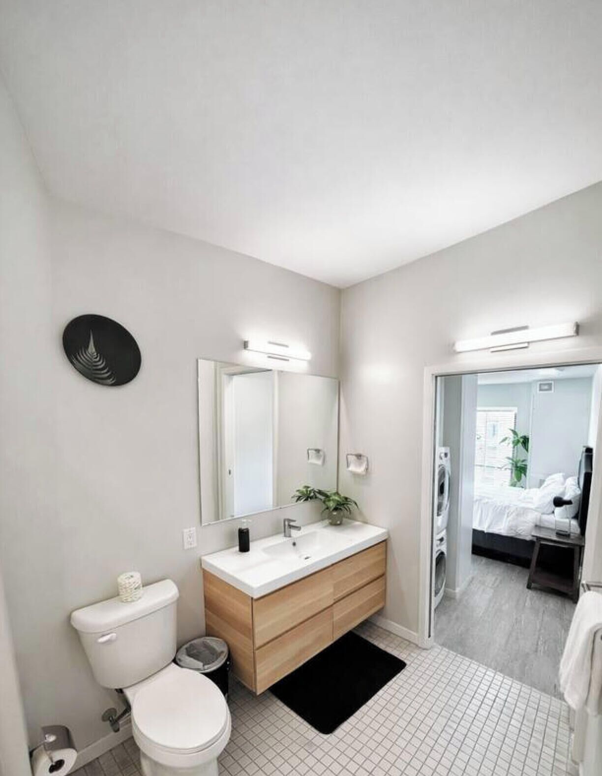 A modern bathroom with a toilet, sink, and mirror. There is a black rug on the floor and a plant on the sink.
