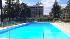 A rectangular swimming pool with a clear blue water, surrounded by a metal fence, trees, and a multi-story building in the background. - Luxurious Vintage 3 Rooms (Antibes)