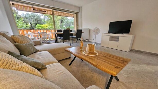 A modern living room with a beige sofa, a wooden coffee table, a TV on a white cabinet, and a balcony with outdoor furniture. - Luxurious Vintage 3 Rooms (Antibes)