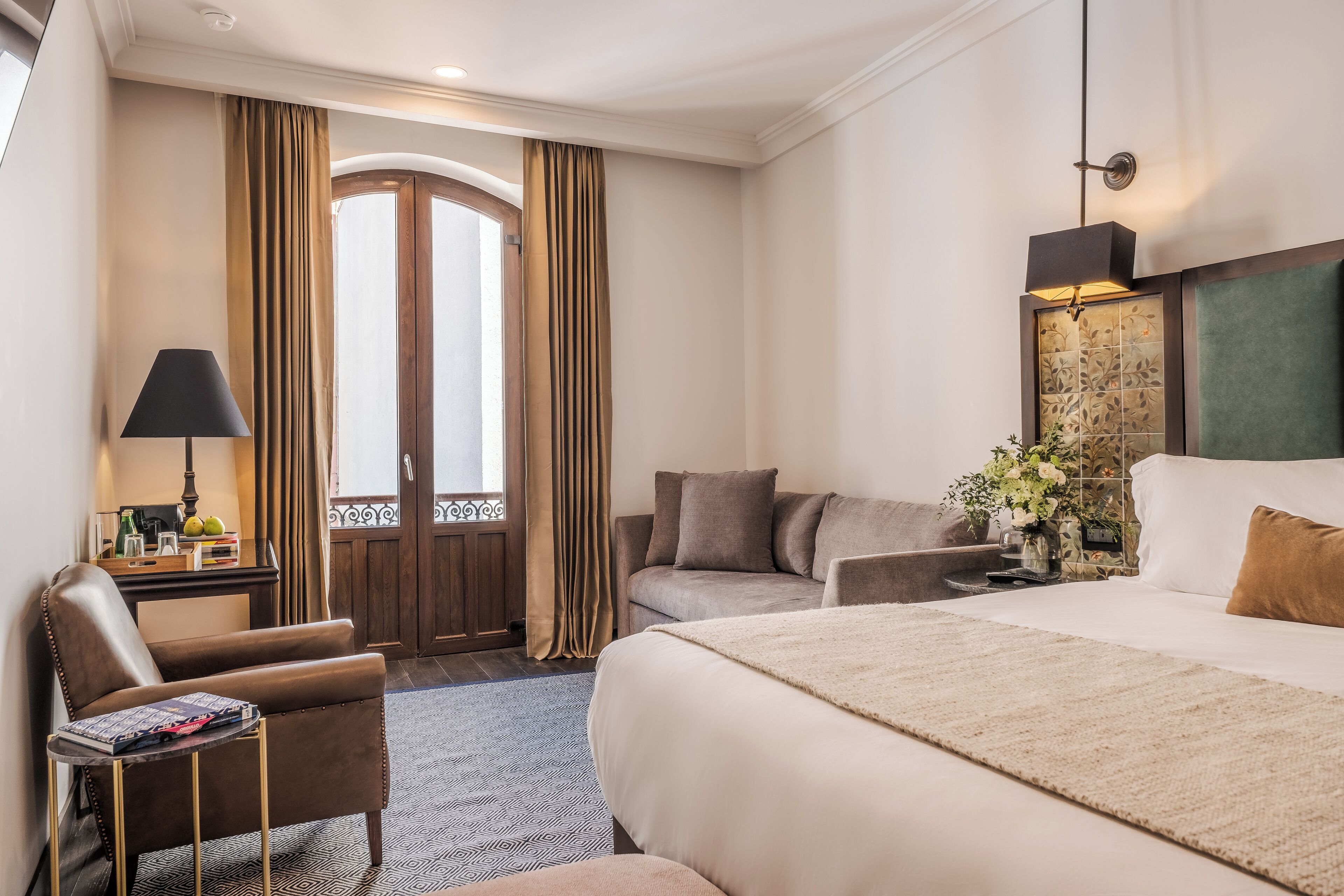 A hotel room with a large bed, a sofa, a chair, a desk with a lamp, and a window with curtains.