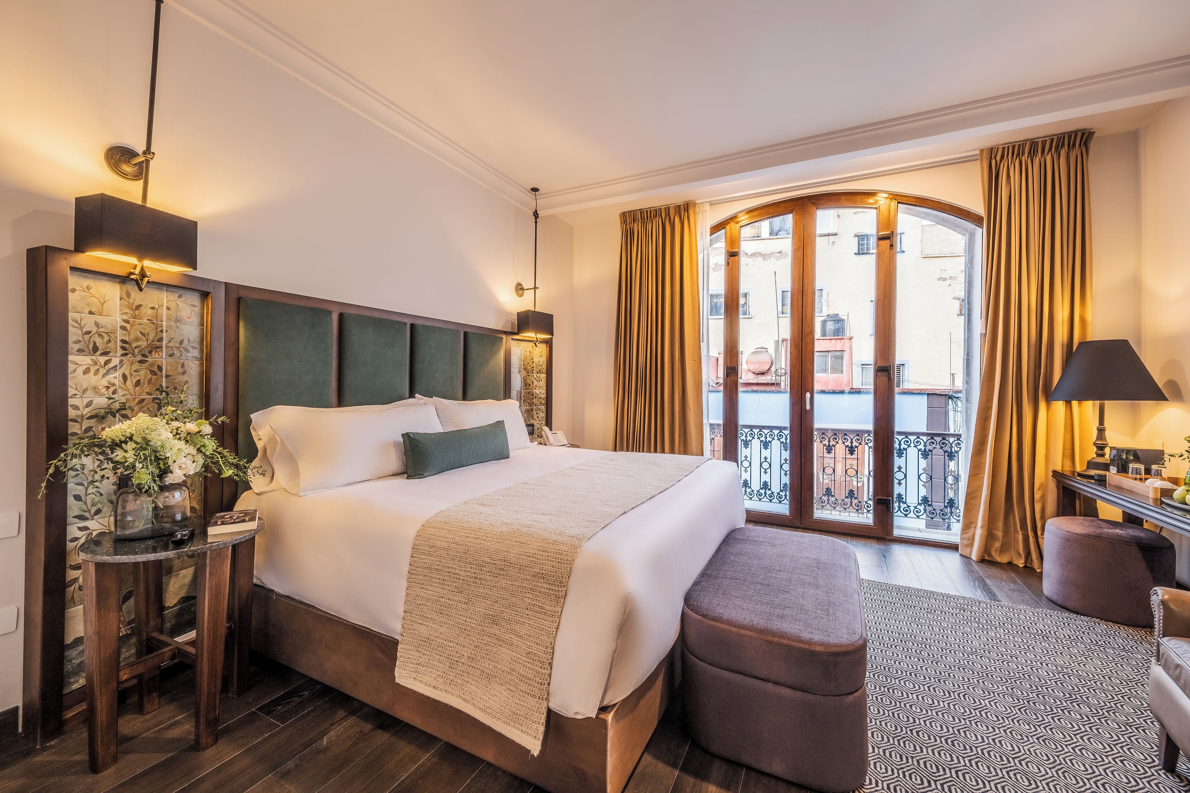 A hotel room with a large bed, a small table with a vase of flowers, a bench, and a view of the city through a glass door.
