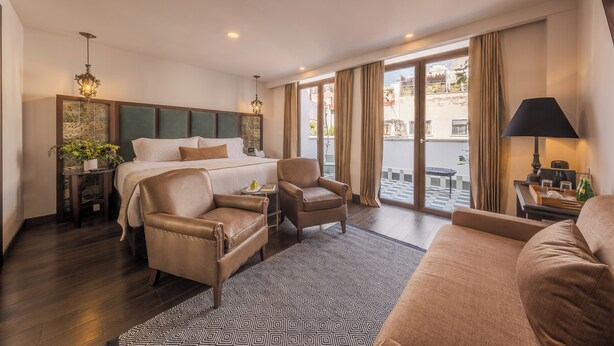 A hotel room with a large bed, two leather armchairs, a sofa, a patterned rug, and a view of the city through large windows. - Casa Kloster Hotel Boutique (Guanajuato)