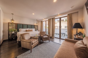 Habitación de hotel con una cama grande, dos sillones de cuero, un sofá, un tapete estampado y vistas a la ciudad a través de amplios ventanales.
