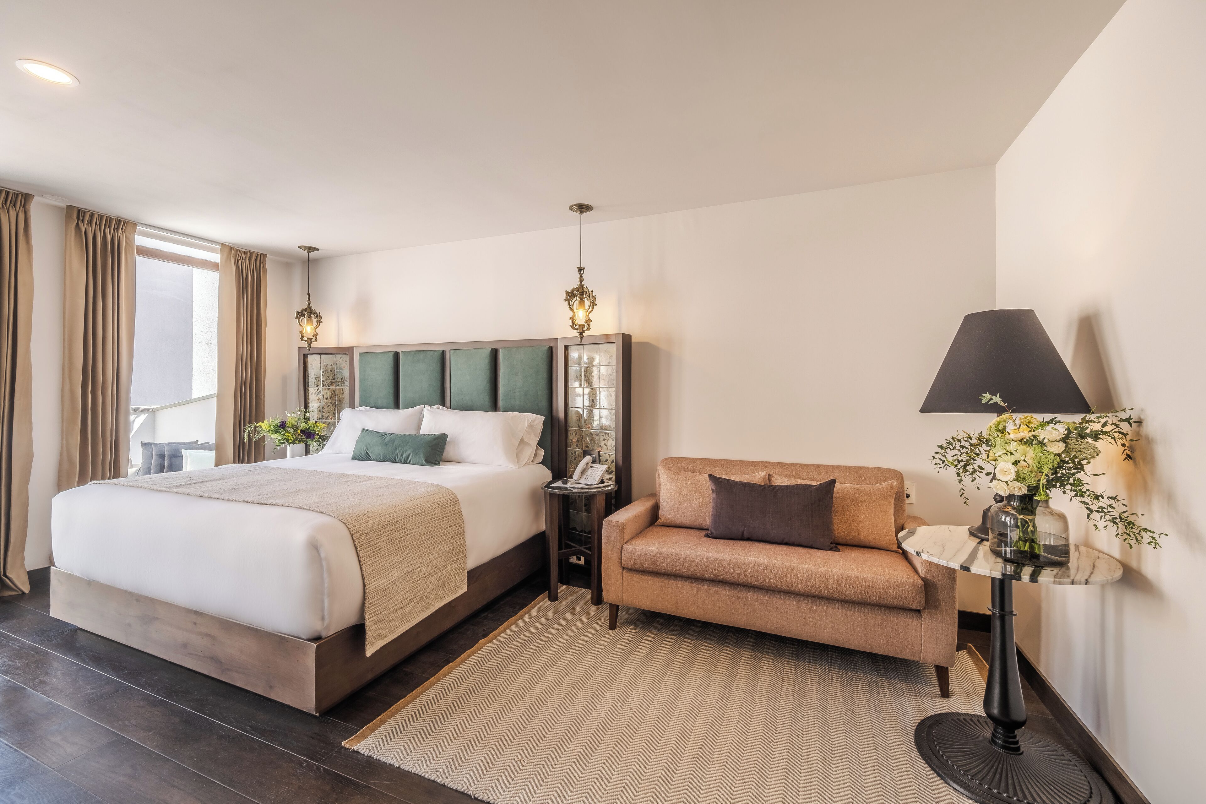 Quarto de hotel moderno com uma cama grande, um sofá, uma mesinha de centro com uma luminária e vista para o exterior.