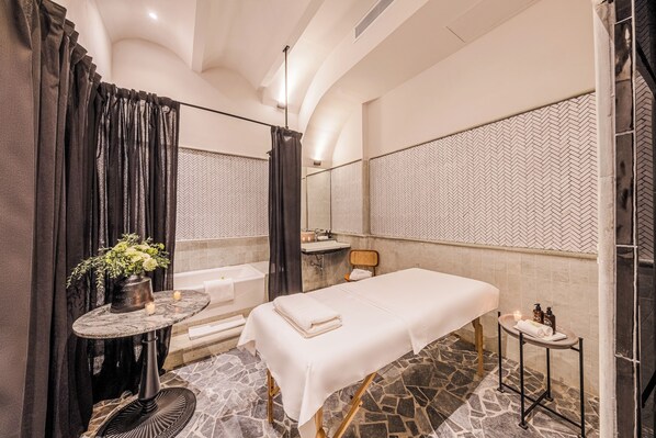 Ein Wellnessraum mit Massageliege, einem kleinen Tisch mit Kerzen, einer Badewanne und einem Spiegel an der Wand.