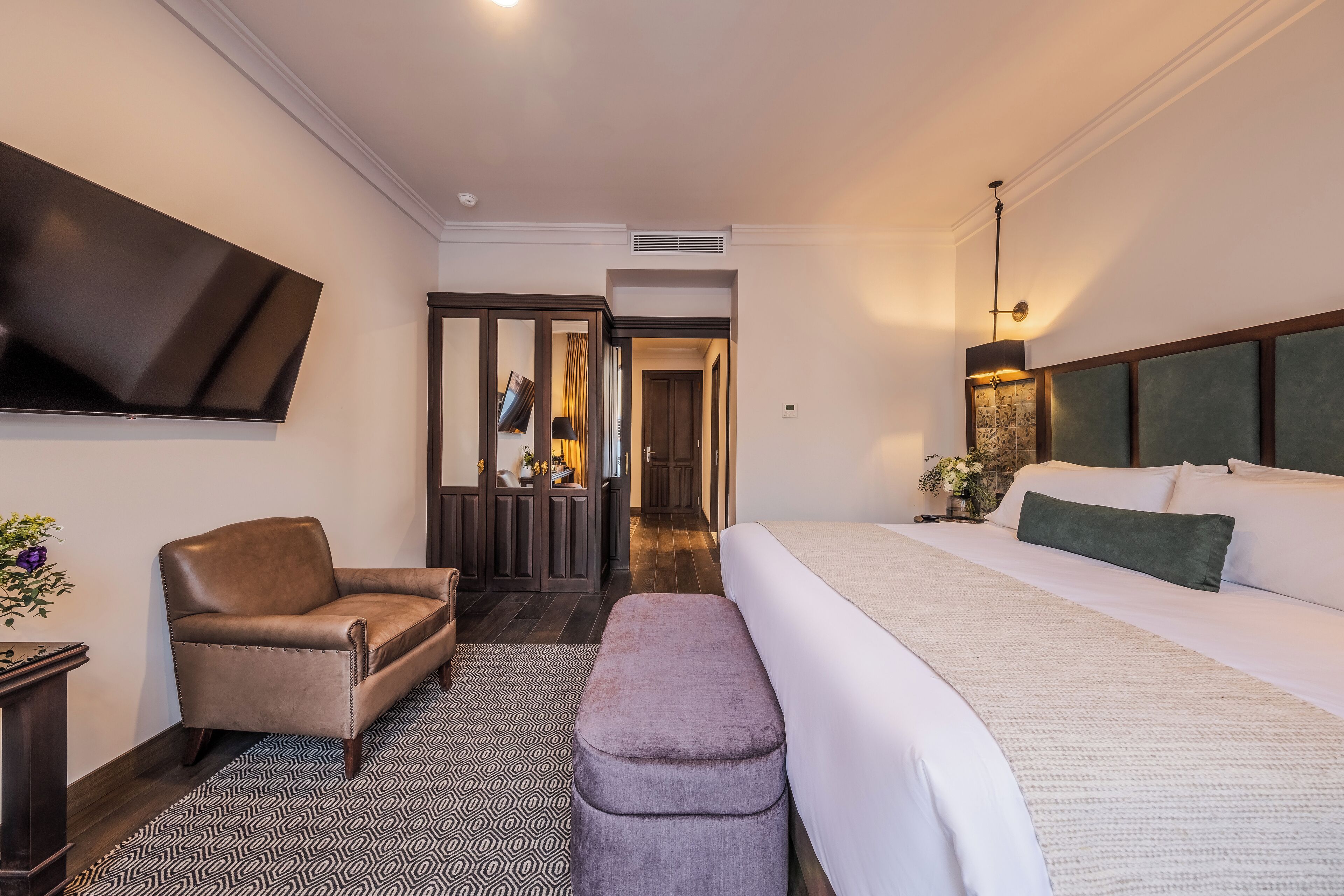 A hotel room with a large bed, a chair, a bench, a TV mounted on the wall, and a hallway leading to another room.