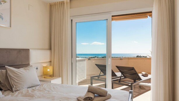 Habitació d'hotel amb llit, una meseta, una làmpada, vista al mar i un balcó amb cadires.